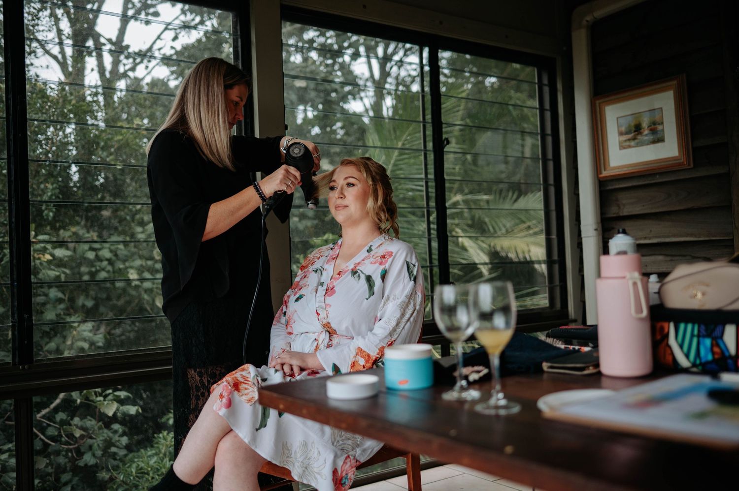 A pre-wedding preparation scene with styling taking place in a sunlit room with wine glasses on the table.