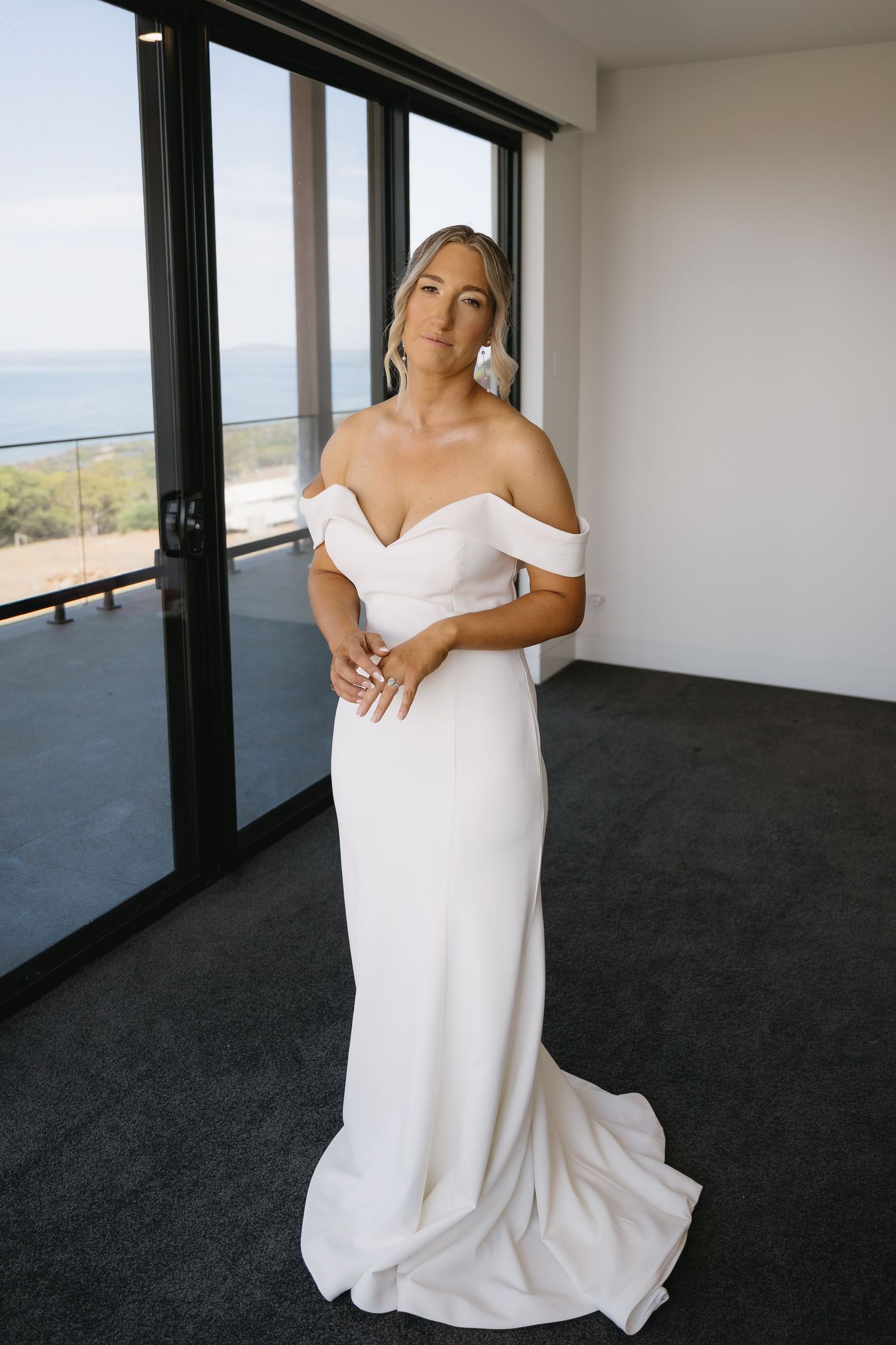 A bride in an elegant off-shoulder white wedding dress poses in a modern room with large windows and scenic views.