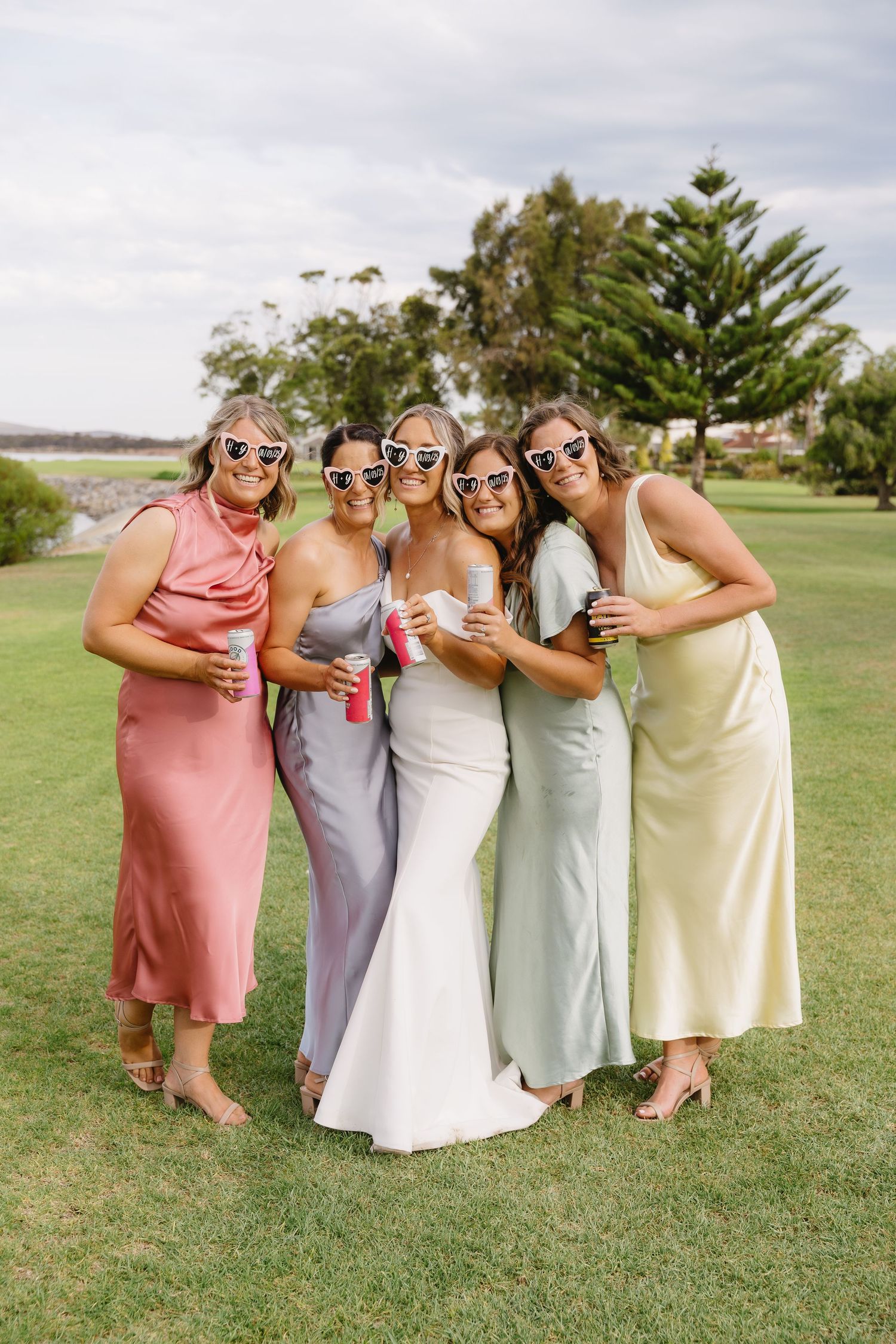 Bridesmaids wearing pastel dresses pose playfully on a grassy lawn.