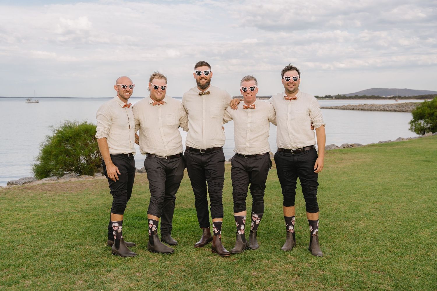 Groomsmen in matching attire pose together on grass with water in the background.