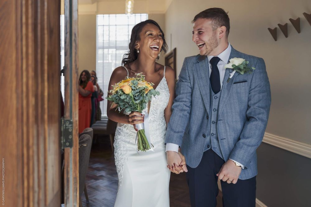 A happy couple laughs together while holding hands at their wedding ceremony in a bright interior space.