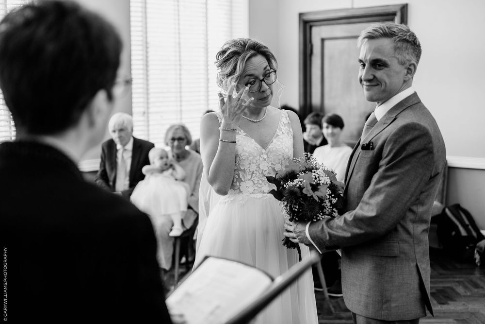 Black and white photo of an emotional bride wiping tears during indoor wedding ceremony while holding bouquet.