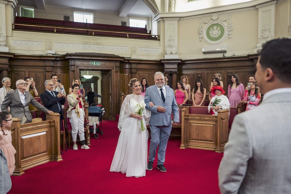 Newlyweds walk down red-carpeted aisle in historic courthouse ceremony room as guests watch from wooden pews.