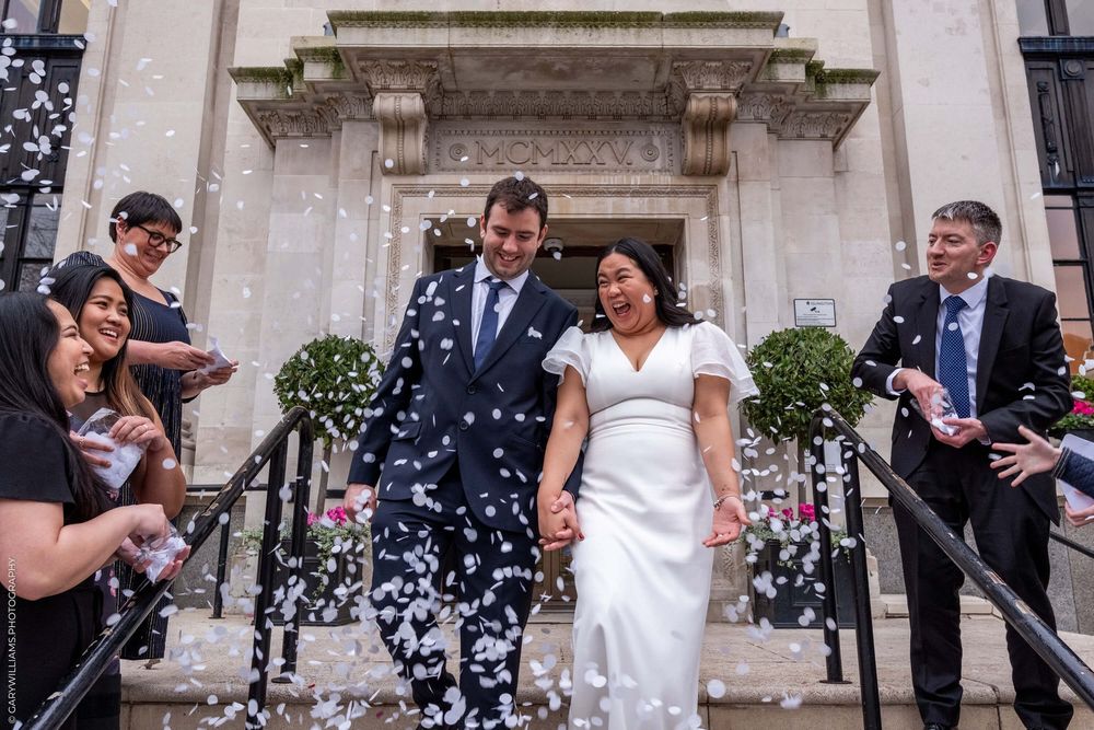 Confetti rains down on couple exiting an elegant building with stone columns and potted plants by the entrance.