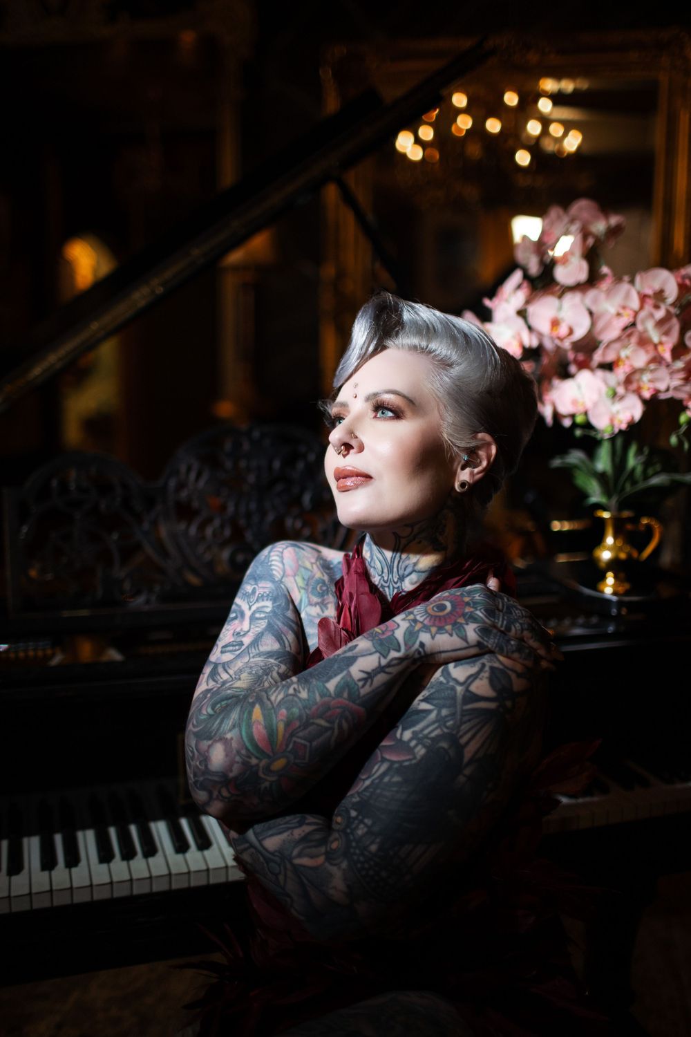 Moody portrait in front of piano with pink flowers, dark ambiance, extensive tattoo artwork visible on arms.