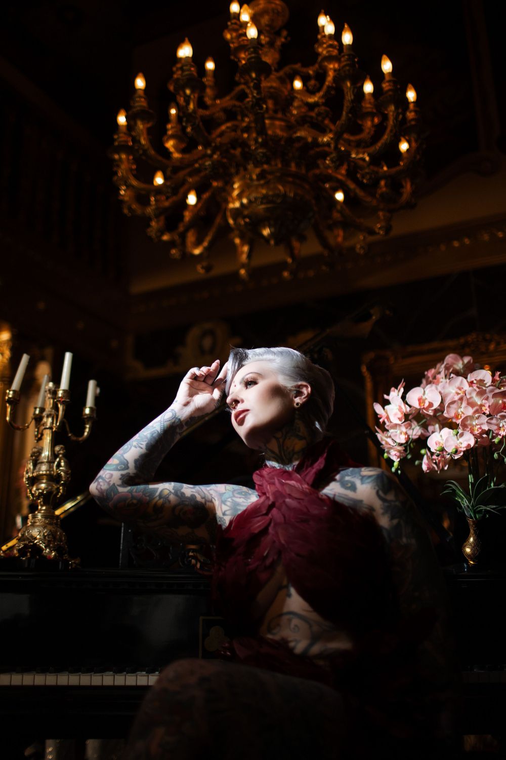 A dramatic portrait in a vintage room with a glowing chandelier, burgundy dress, and pink flowers by a piano.