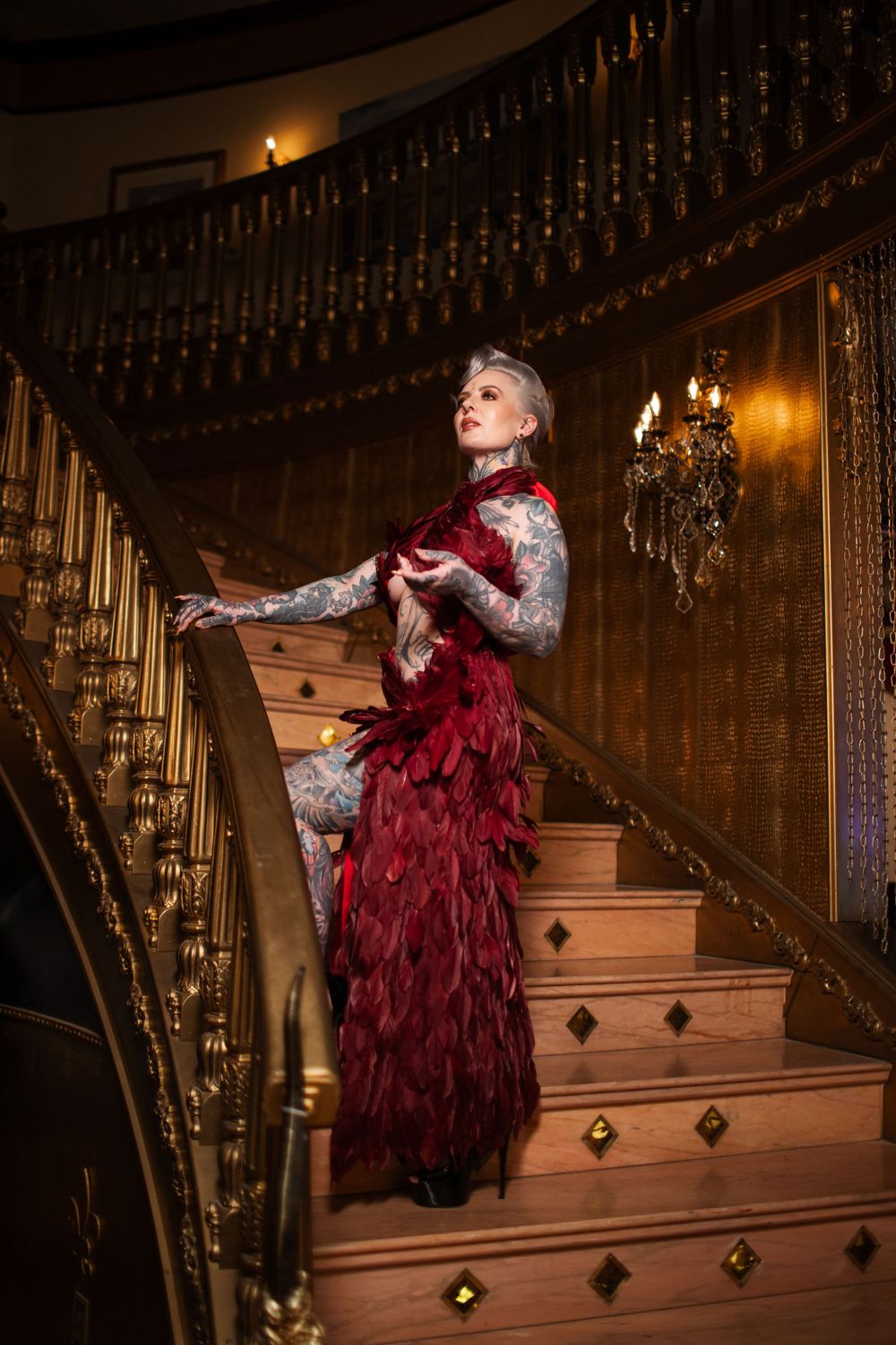 A person in a dramatic red and white Victorian gown poses elegantly on an ornate wooden staircase with chandelier lighting.