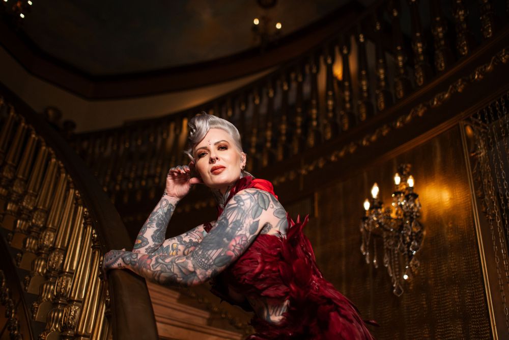Someone in a vintage red and white dress poses dramatically in an ornate room with a grand staircase and chandeliers.