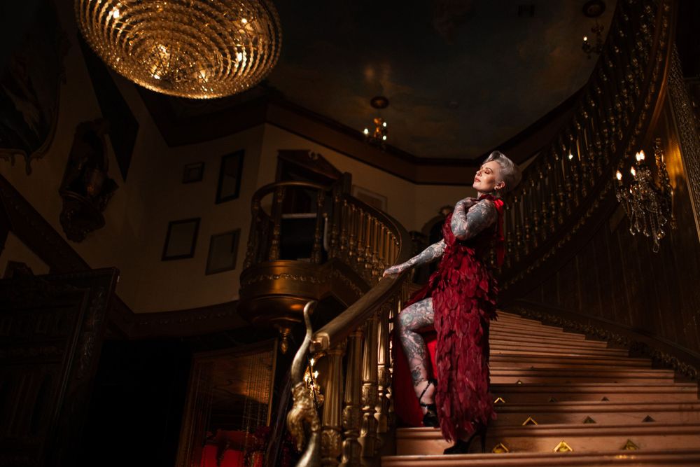 Elegant figure in a sparkly red dress poses dramatically on grand staircase under warm chandelier lighting.