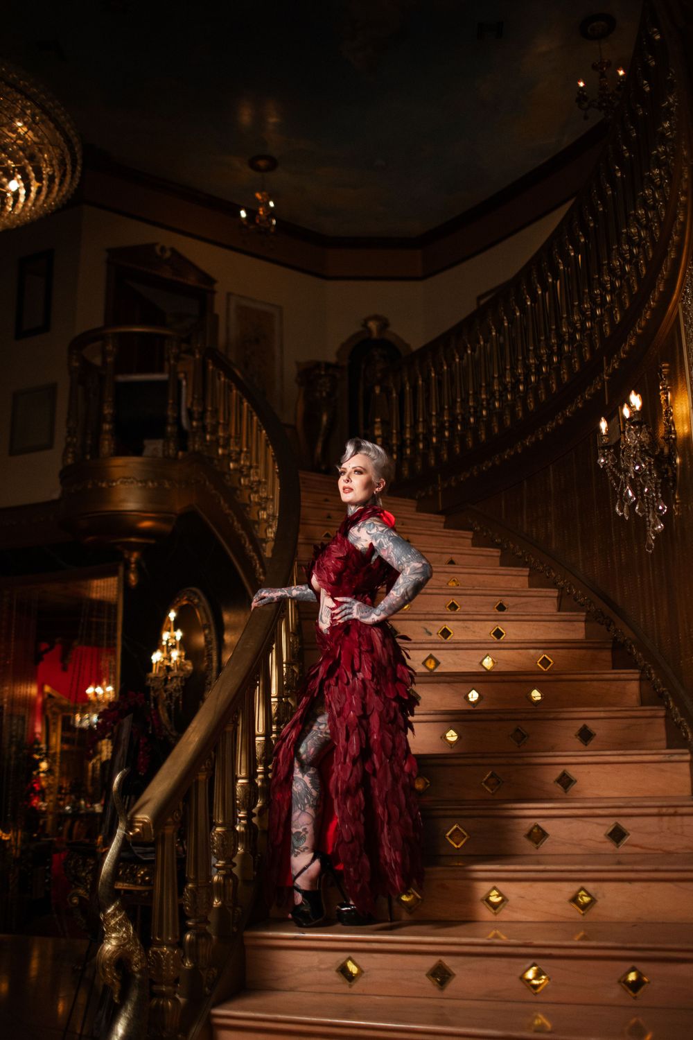 A glamorous figure in a flowing burgundy dress poses on an ornate staircase beneath crystal chandeliers.