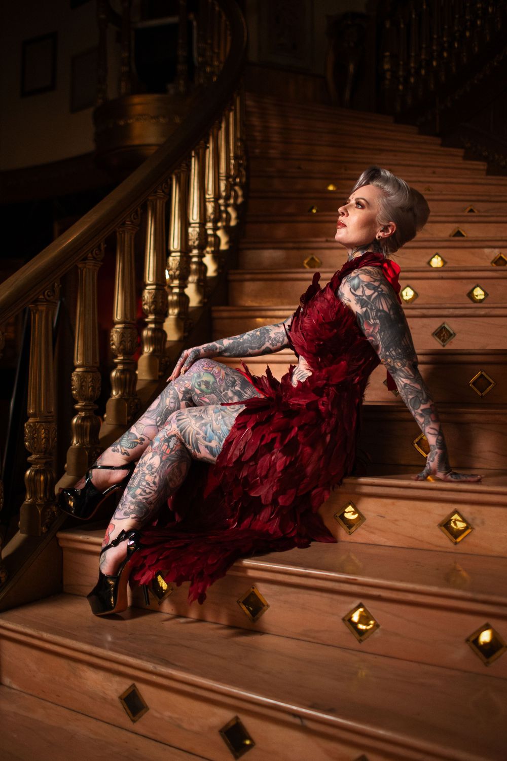Woman in red velvet dress and tattoos posing dramatically on vintage wooden staircase with decorative details.