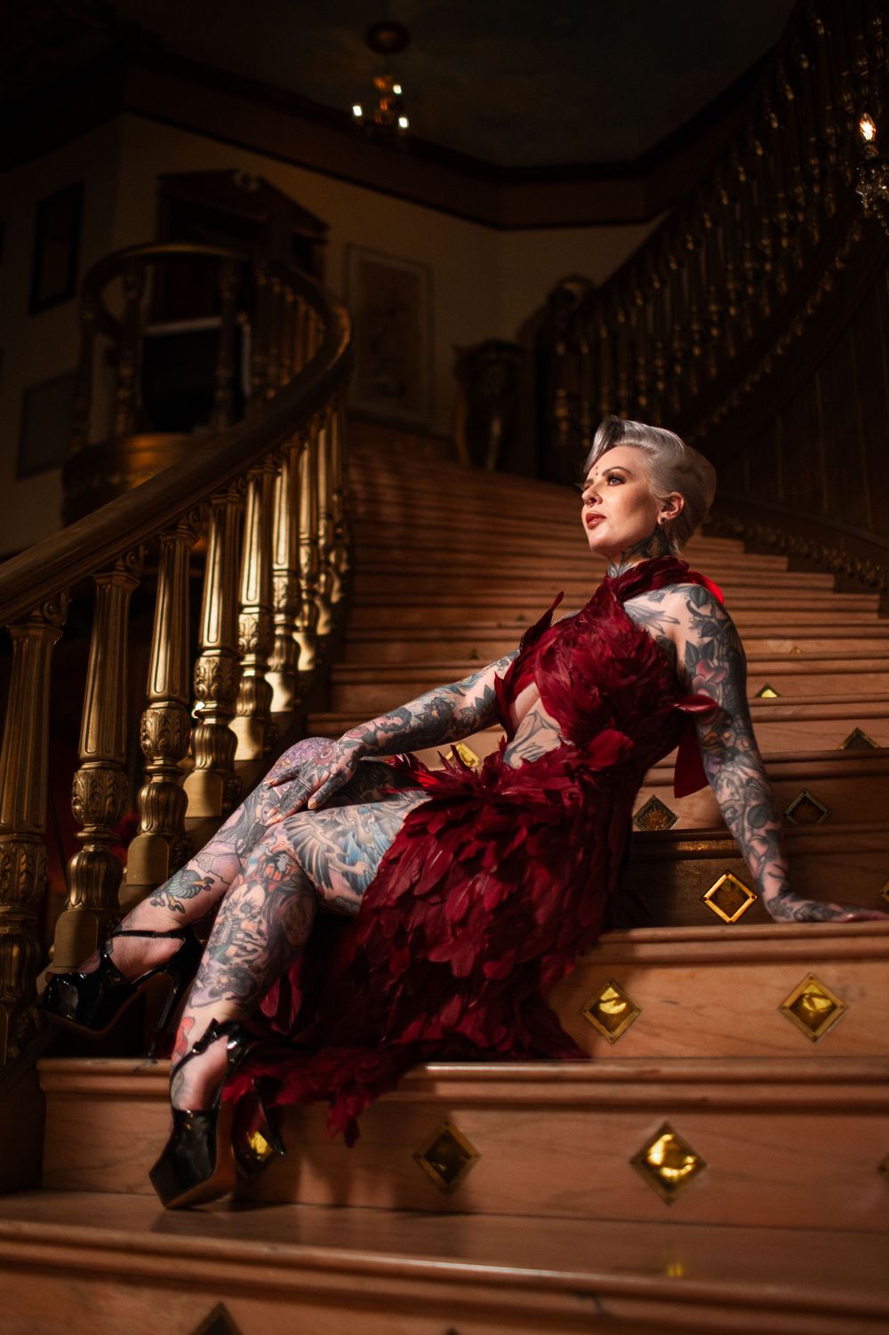 A person in a red velvet dress and tattoos poses dramatically on wooden stairs with ornate gold railings.