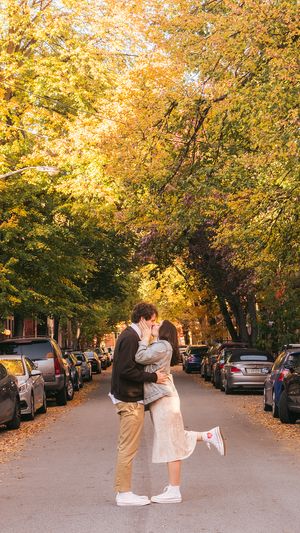 A romantic autumn street scene with yellow trees and parked cars as a couple embraces on the road.