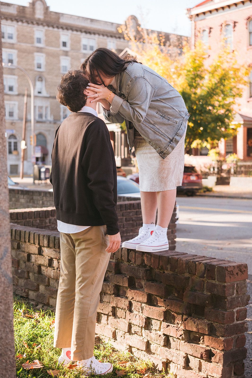 A couple shares a romantic moment on a brick wall with historic buildings and fall foliage in the background.