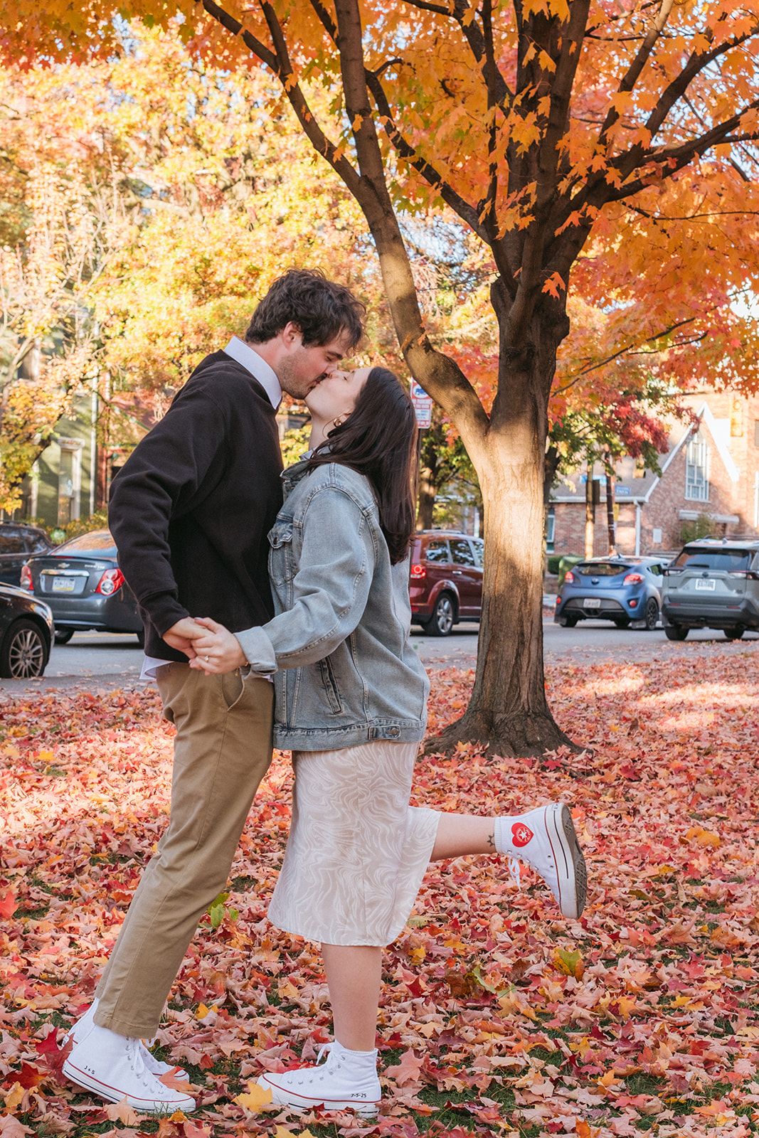 A romantic couple poses playfully among fallen autumn leaves with vibrant orange foliage behind them.