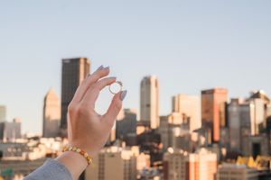 Hand holds an engagement ring against a city skyline backdrop at sunset.