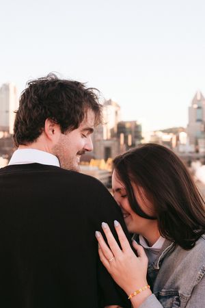 A romantic couple embraces against a sunset cityscape backdrop, with the woman resting her hand on her partner's shoulder.