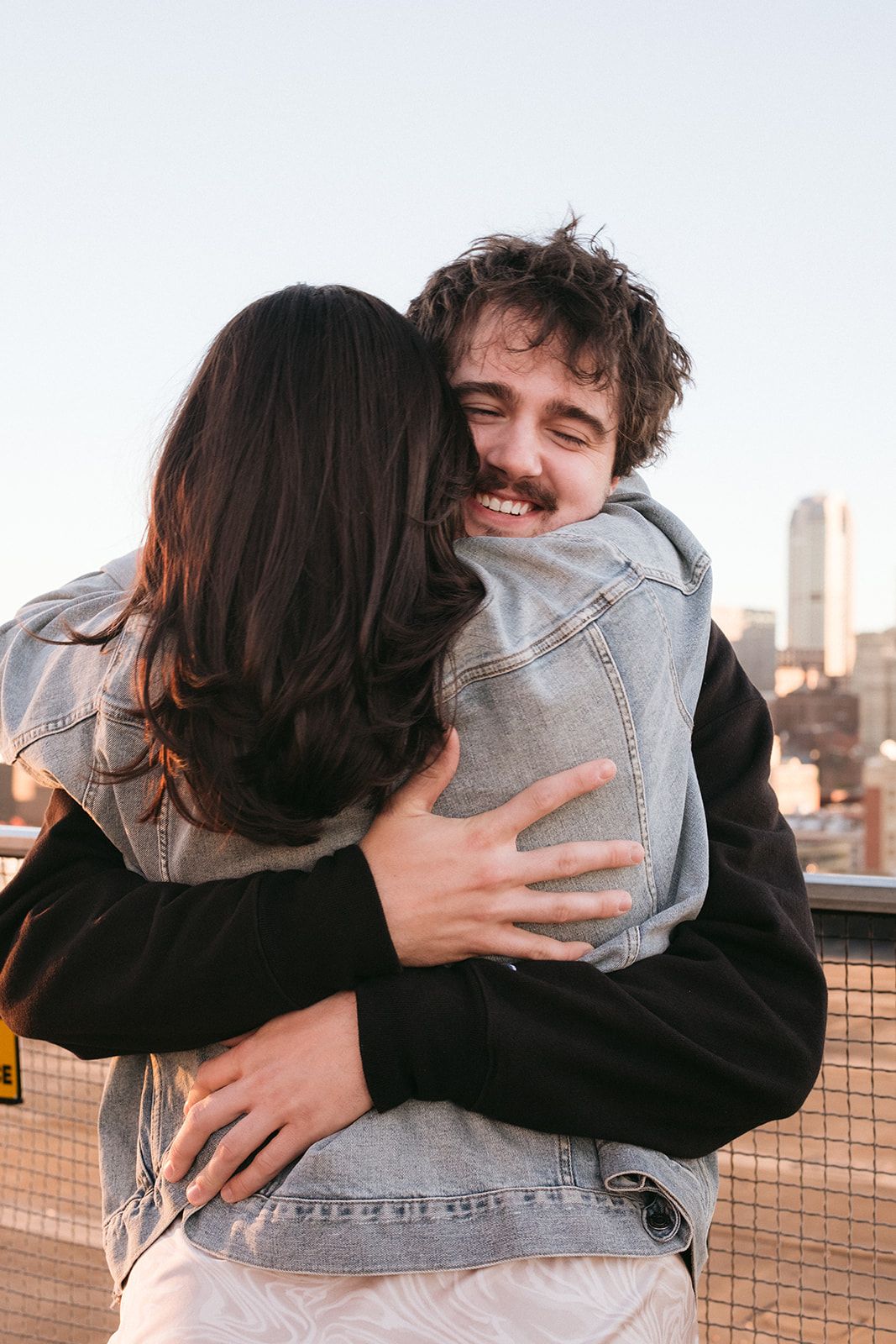 Two people share a warm embrace on a rooftop with urban buildings in the background.