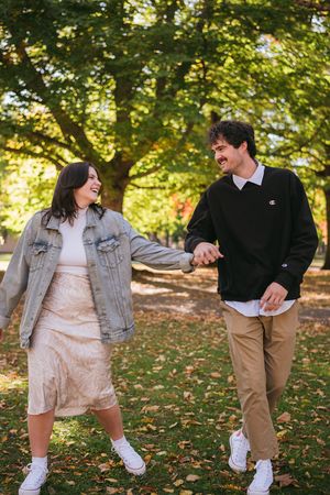 A couple holding hands and laughing together in a park during autumn with fallen leaves on the ground.