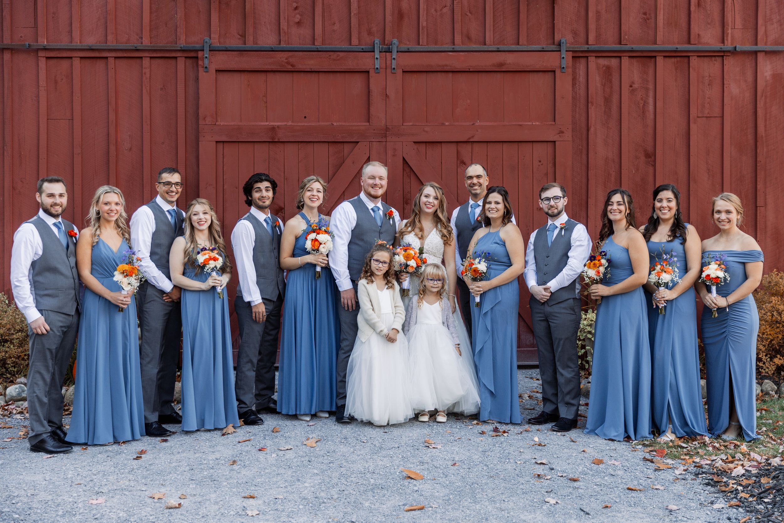 Wedding party in coordinated blue dresses and grey suits poses in front of red barn.