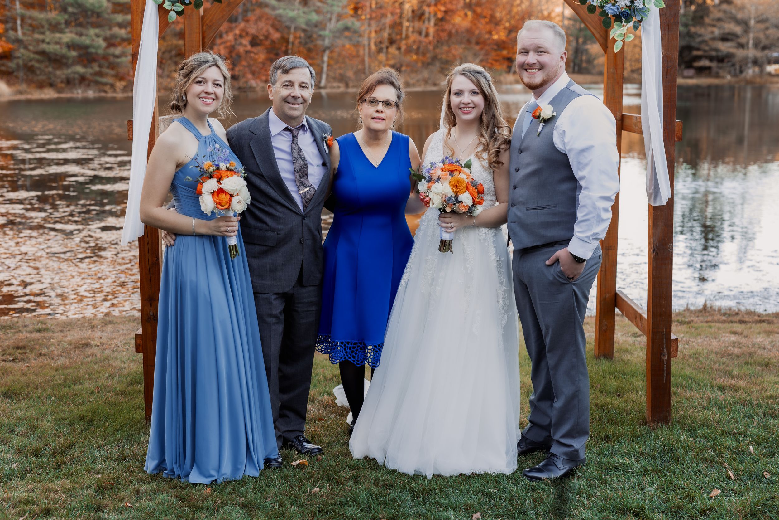 Small wedding party poses together under wooden arch by lakeside during autumn ceremony.