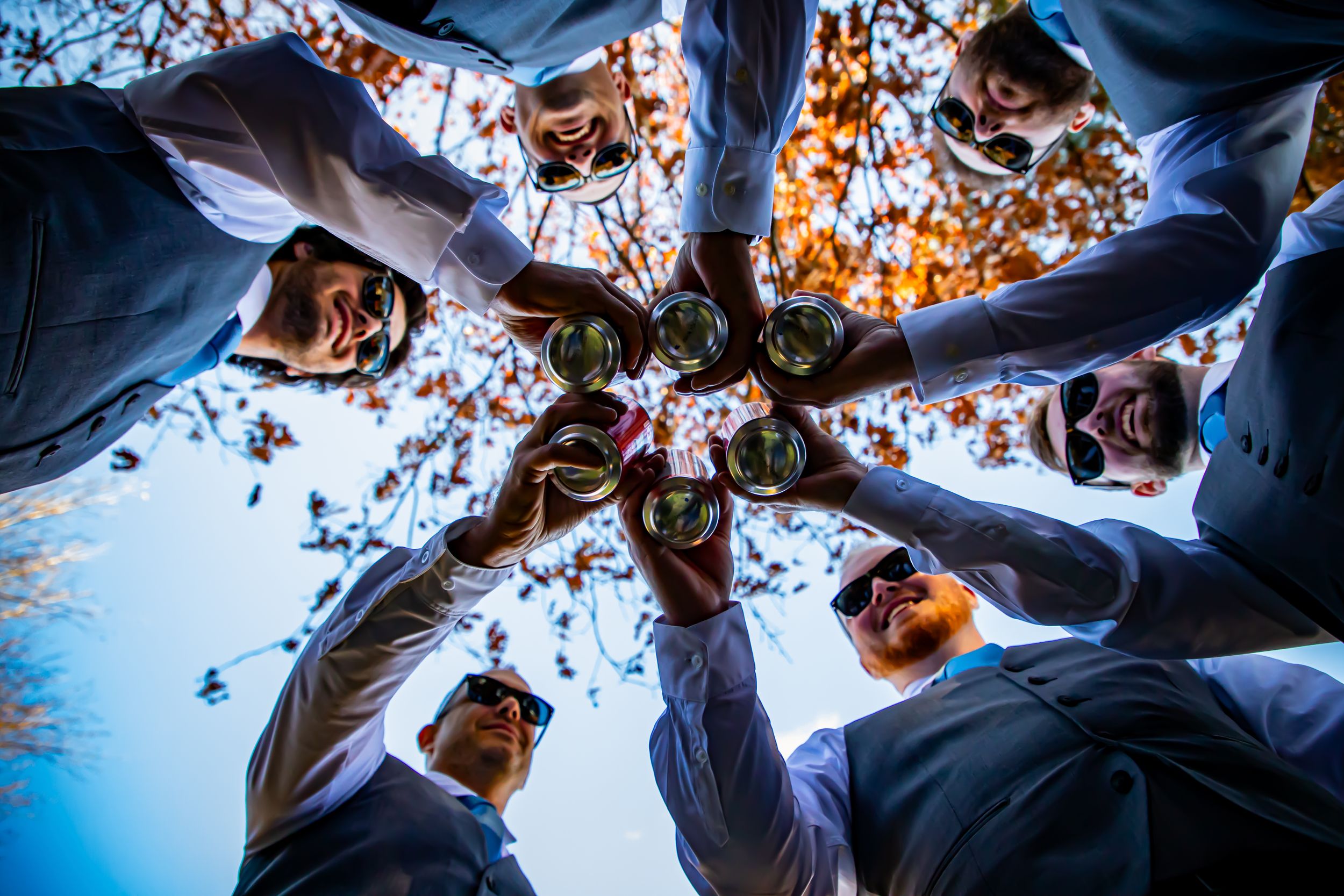 Groomsmen toast with drinks in creative low-angle circle shot against autumn sky.