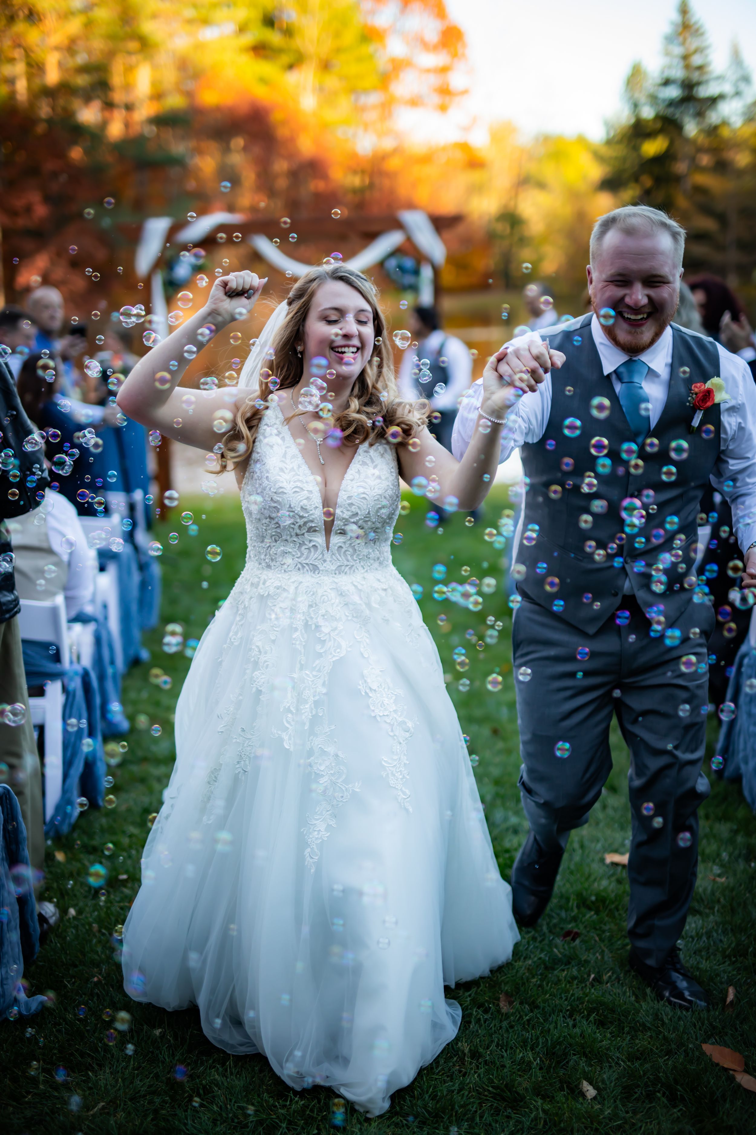Newlyweds walk down aisle surrounded by bubbles during outdoor autumn wedding ceremony.