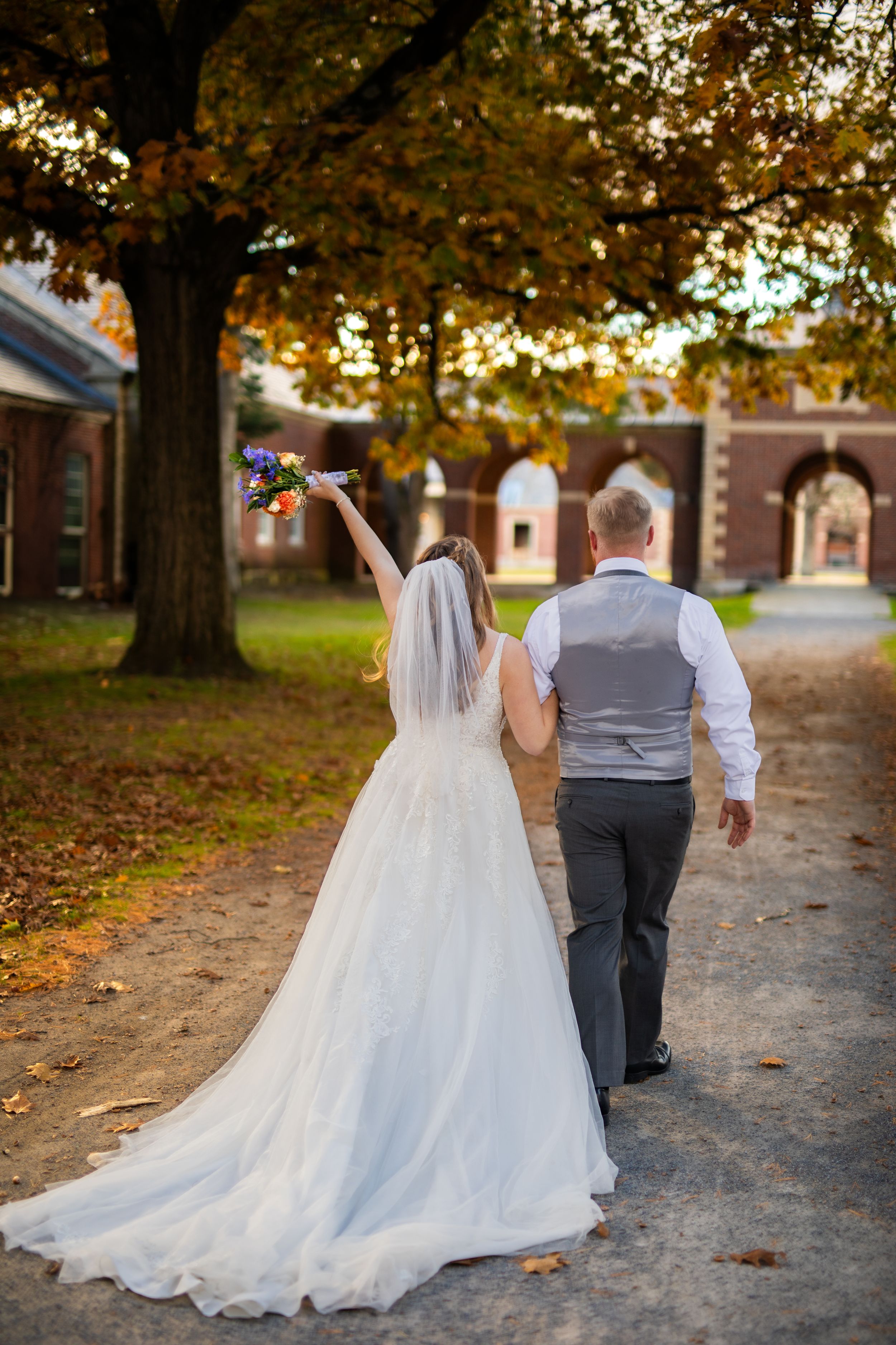 Couple walks hand in hand on autumn path with falling leaves and historic buildings.
