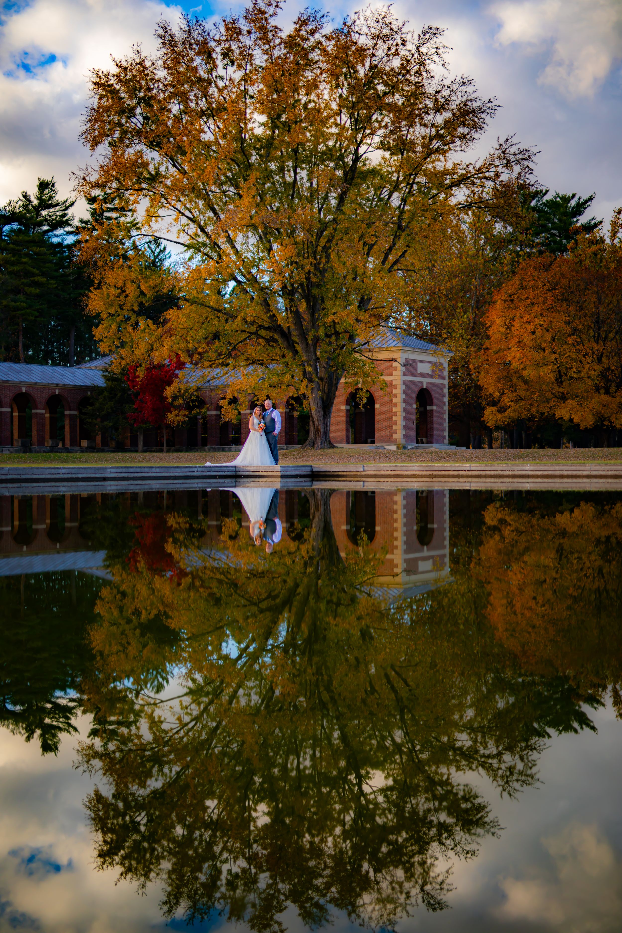 Stunning fall wedding photo reflected in still water with golden trees and brick architecture.