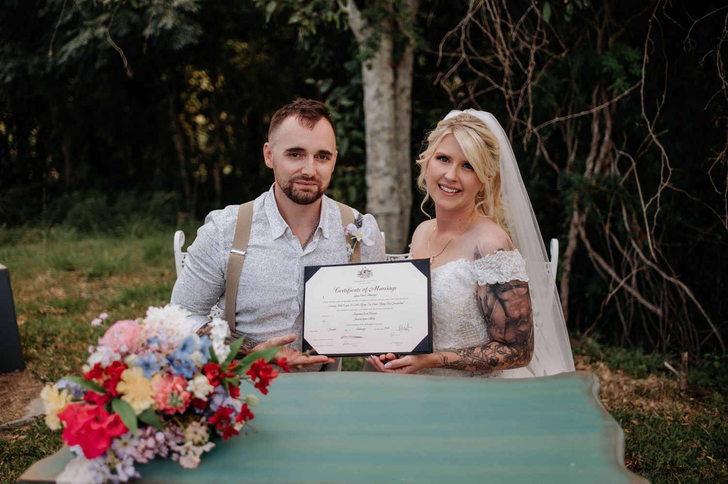 Newlyweds display their marriage certificate at outdoor ceremony with colorful floral arrangement.