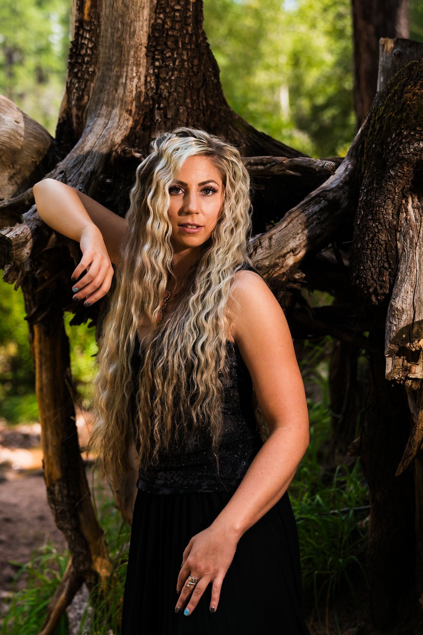 A person with long wavy blonde hair wearing a black dress poses between dark tree trunks in a forest setting.