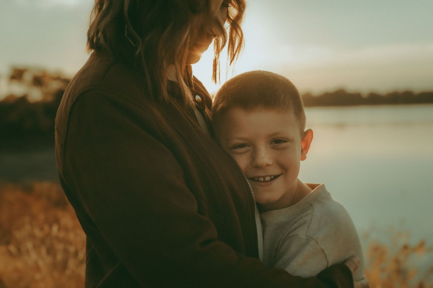 Smiling boy wrapped in mother’s arms at golden hour – authentic motherhood session near the lake in Kansas