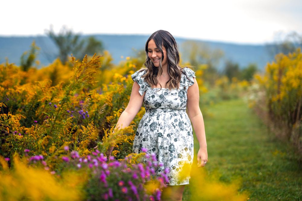 A person in a floral dress walks through a colorful garden with yellow and purple wildflowers against distant mountains.