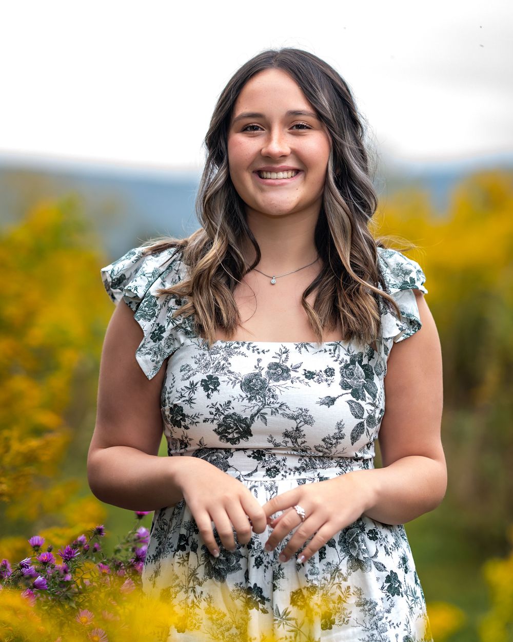 A person in a floral dress poses outdoors among yellow wildflowers and blurred mountains in the background.