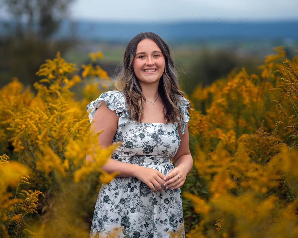 A person in a floral dress poses in a field of vibrant yellow wildflowers with blurred blue sky in background.