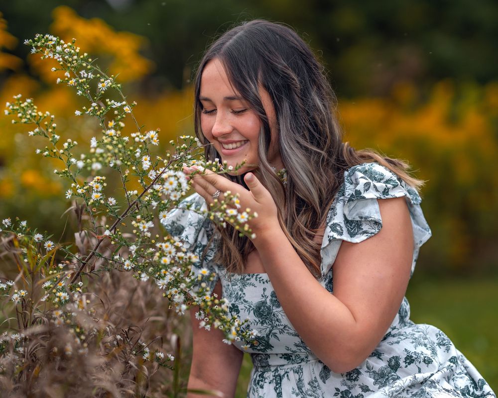 Person in floral dress smells delicate white wildflowers in a garden with yellow flowers in the background.
