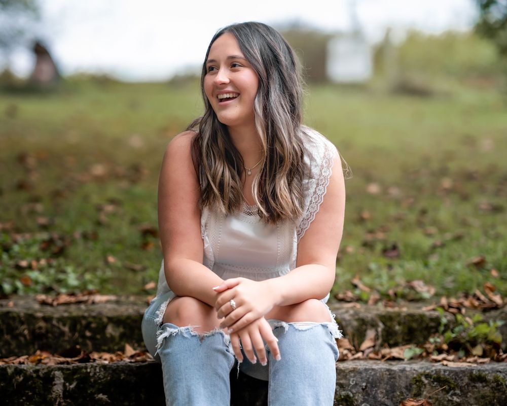 A young woman in a white top and jeans sits on the ground laughing outdoors among scattered autumn leaves.