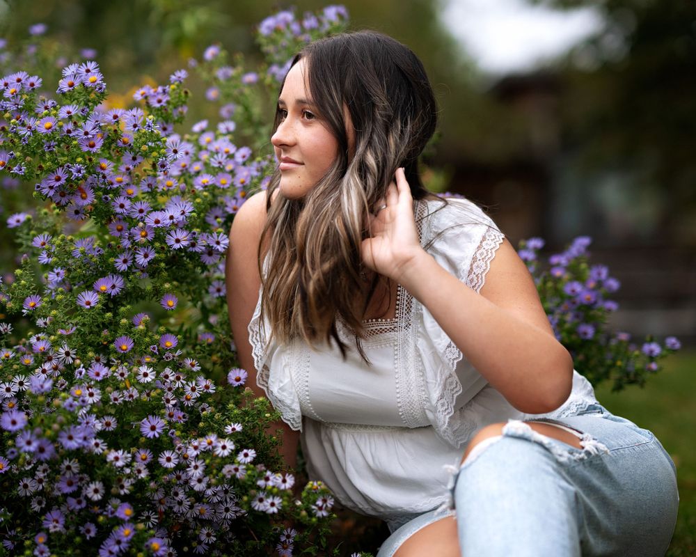 A woman in a white lace top poses beside blooming purple flowers in a garden setting.