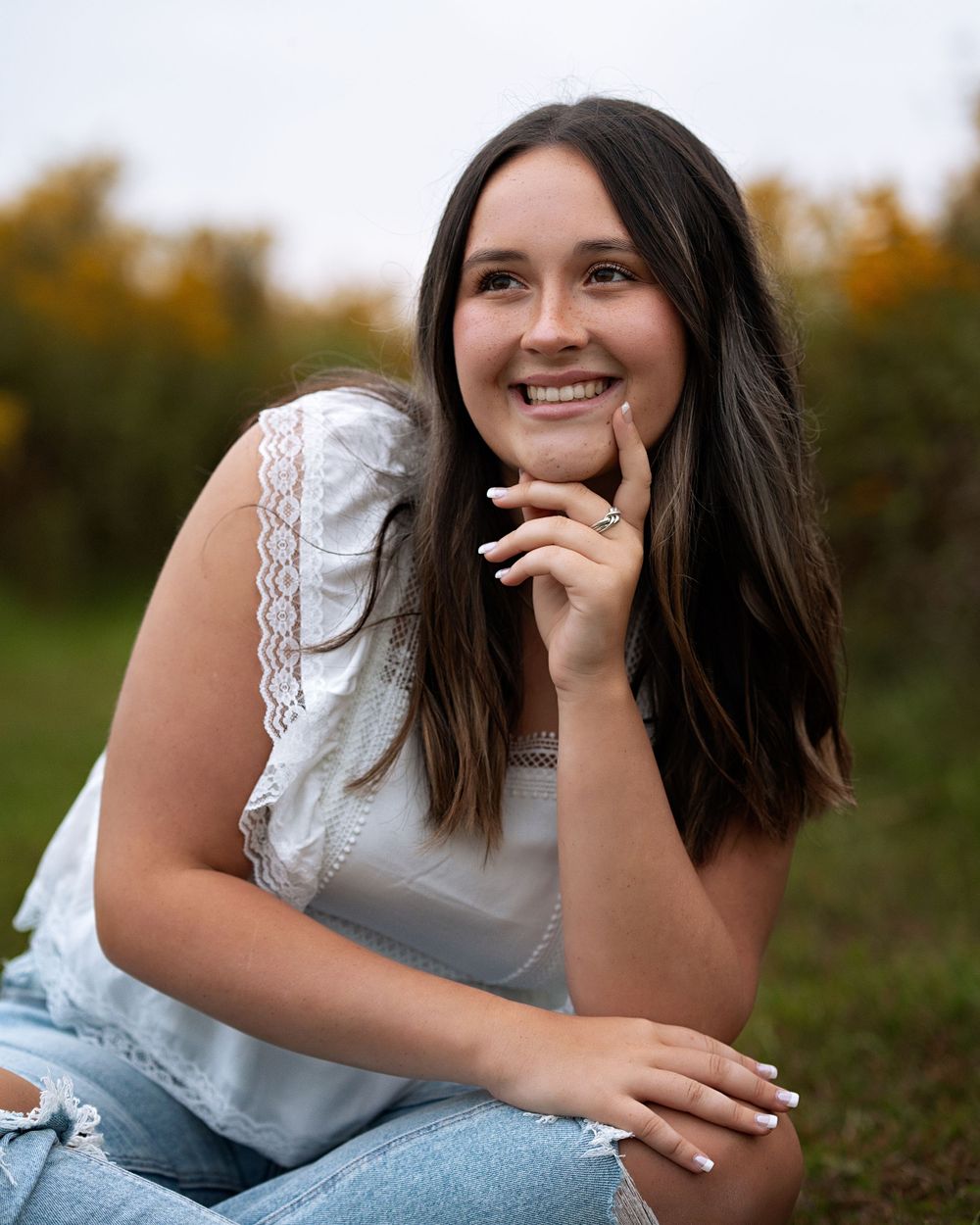 A woman in a white top and jeans sits outdoors with an autumn background while touching her face.