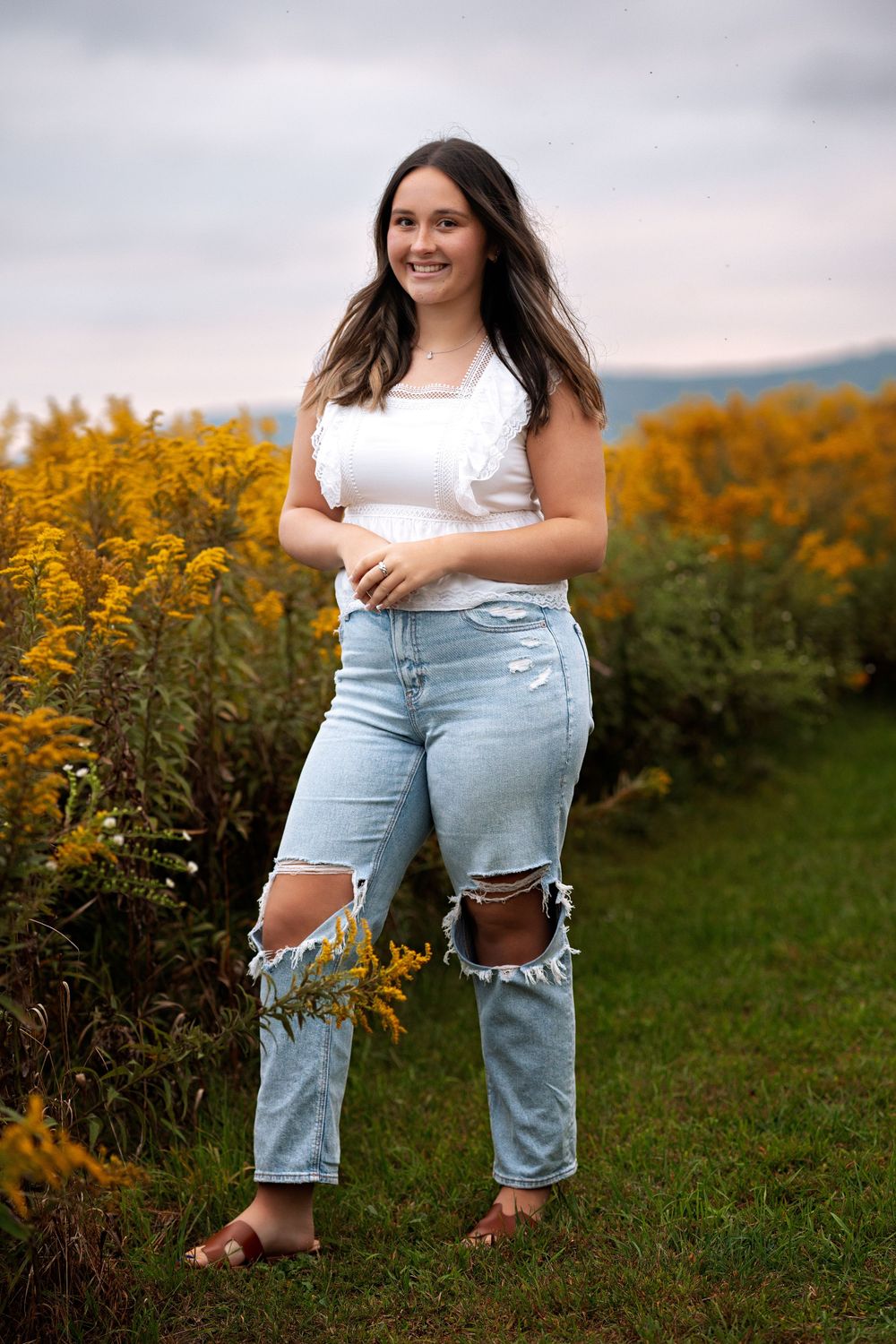 Someone poses in ripped blue jeans and white top against a backdrop of golden wildflowers on a cloudy day.