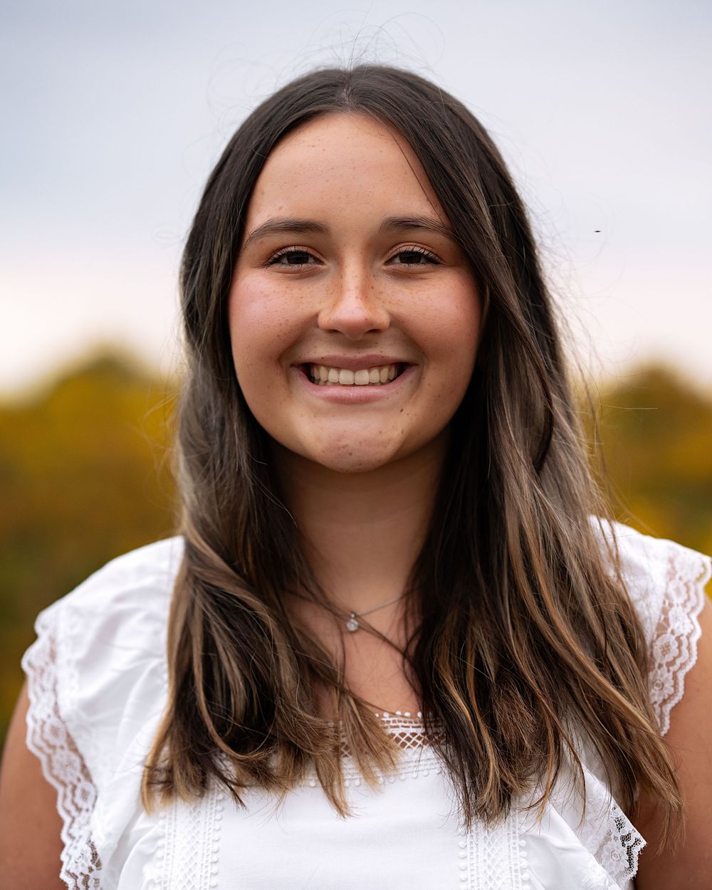 A portrait of someone in a white lace top smiling warmly against an autumn background.