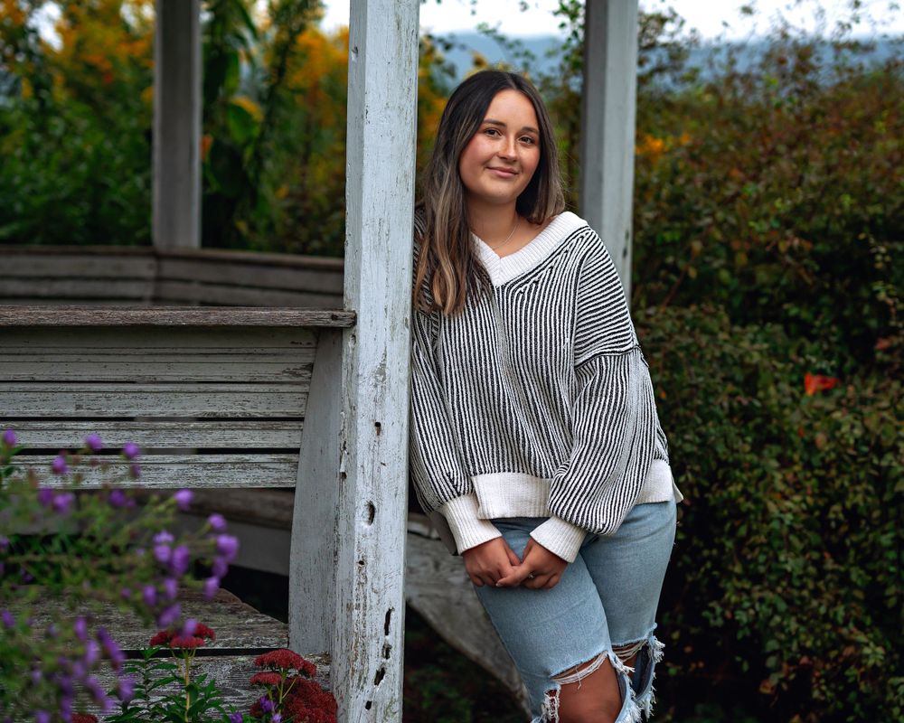A person in a striped sweater and ripped jeans sits casually on wooden steps surrounded by autumn flowers.