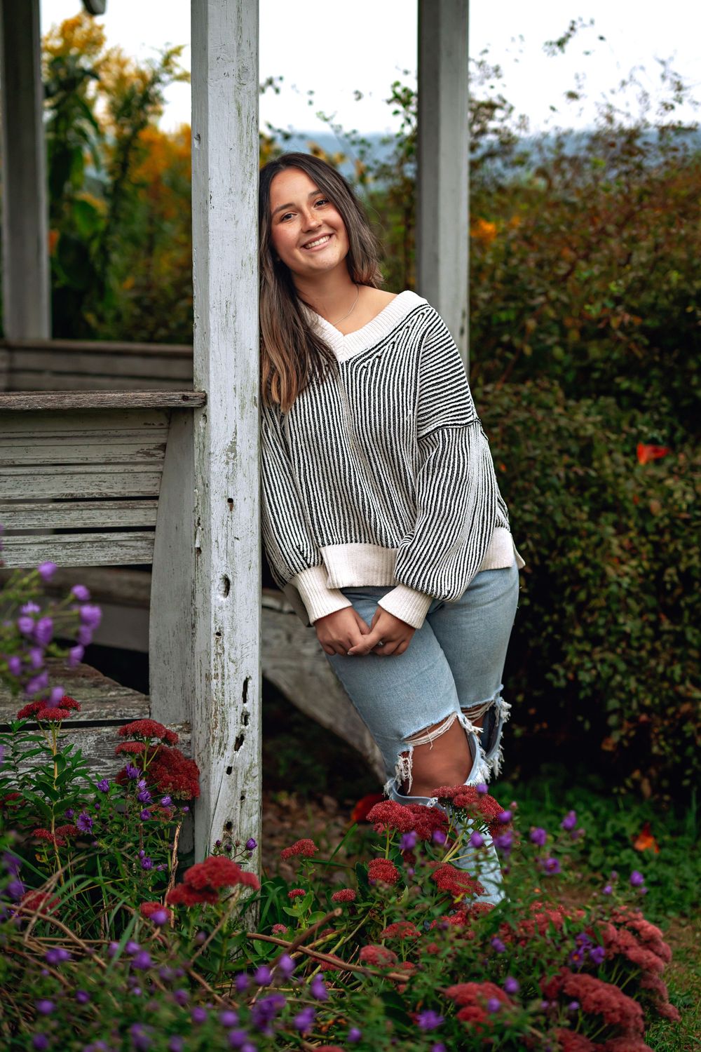 Portrait of someone wearing a striped sweater and distressed jeans standing by a porch with red and purple flowers.