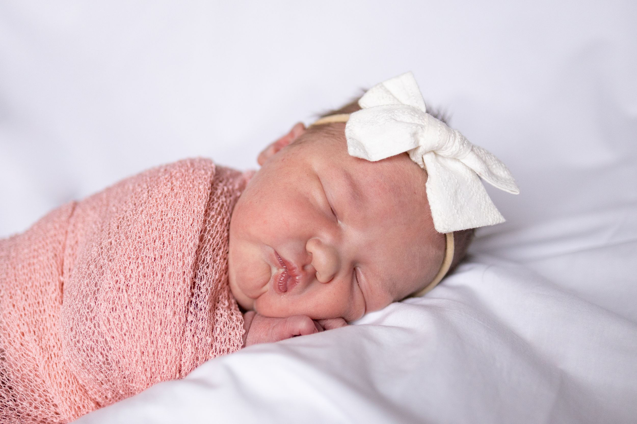 A sleeping newborn baby wrapped in a pink swaddle blanket with a white bow headband resting on white bedding.
Newborn Pose.