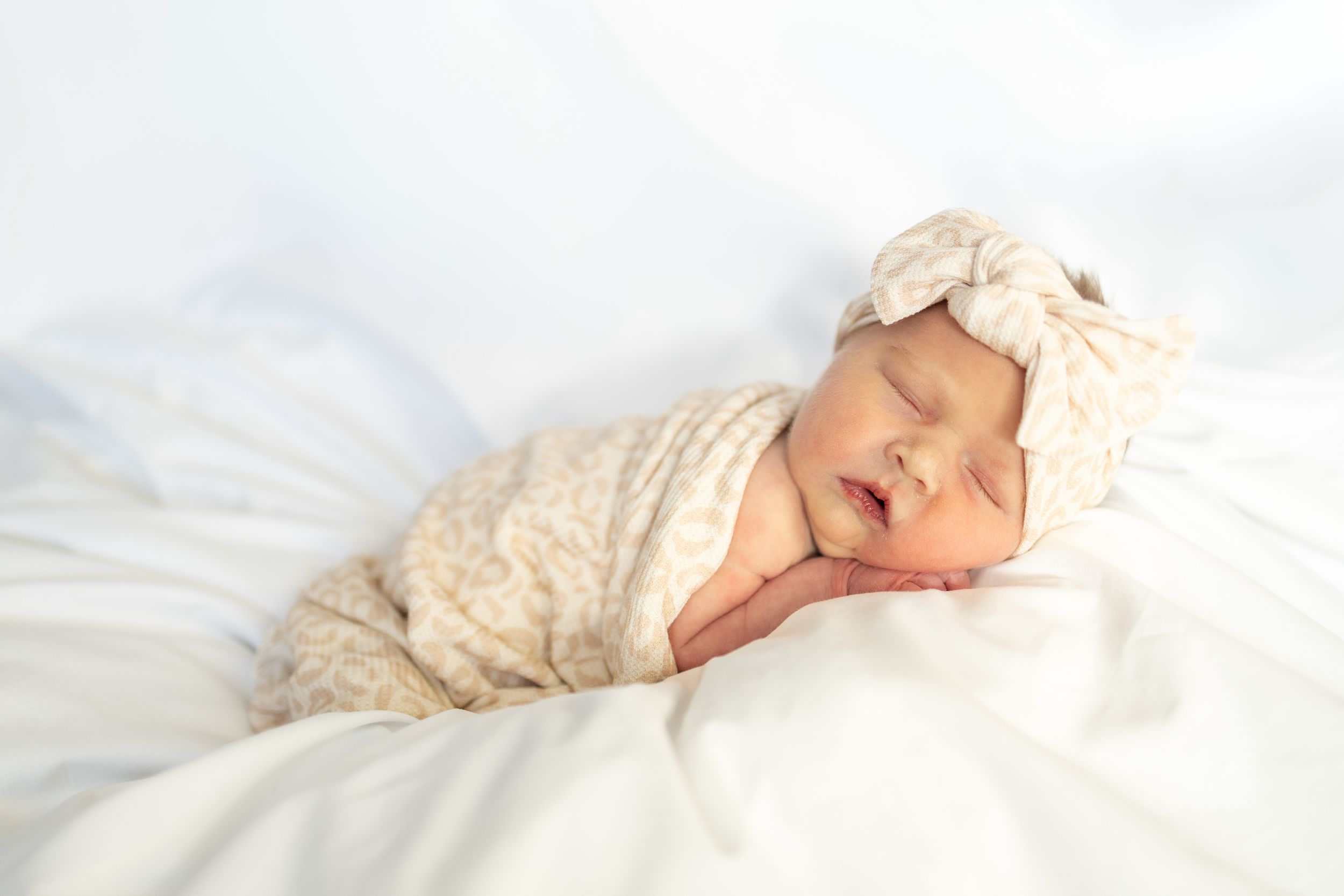 Sleeping newborn baby wrapped in soft white blankets lying on white bedding in natural light.