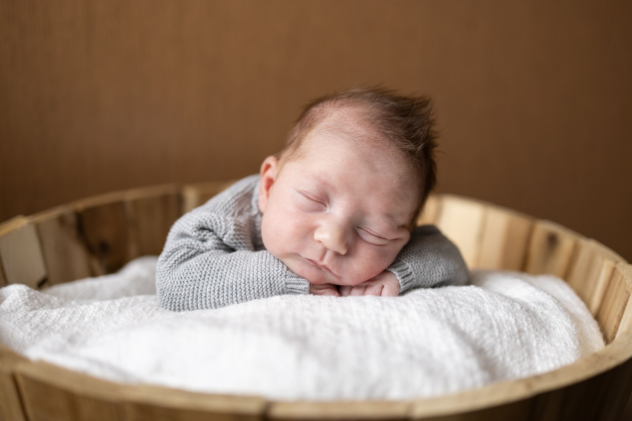 Peaceful sleeping newborn in gray sweater rests on white blanket in wooden bowl.