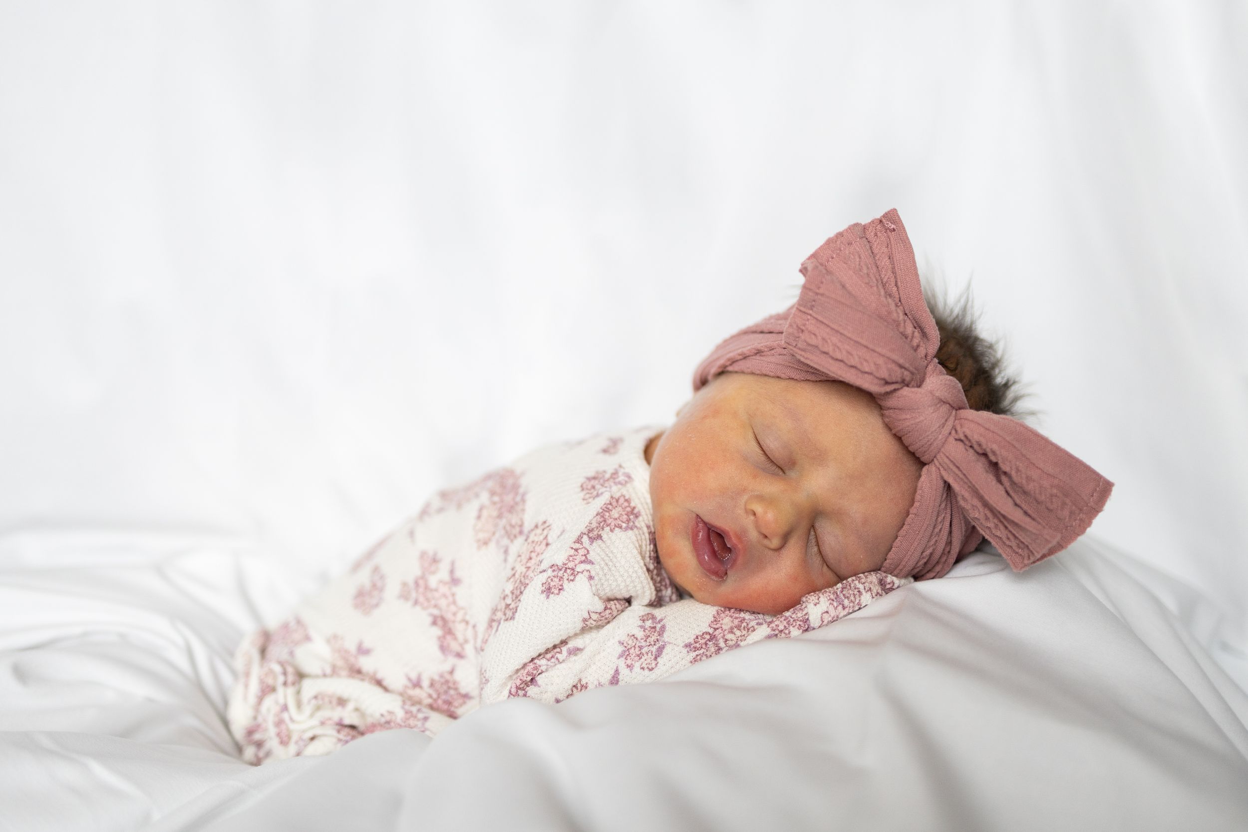 Sleeping newborn baby wearing pink bow headband and floral outfit rests peacefully on white bedding.