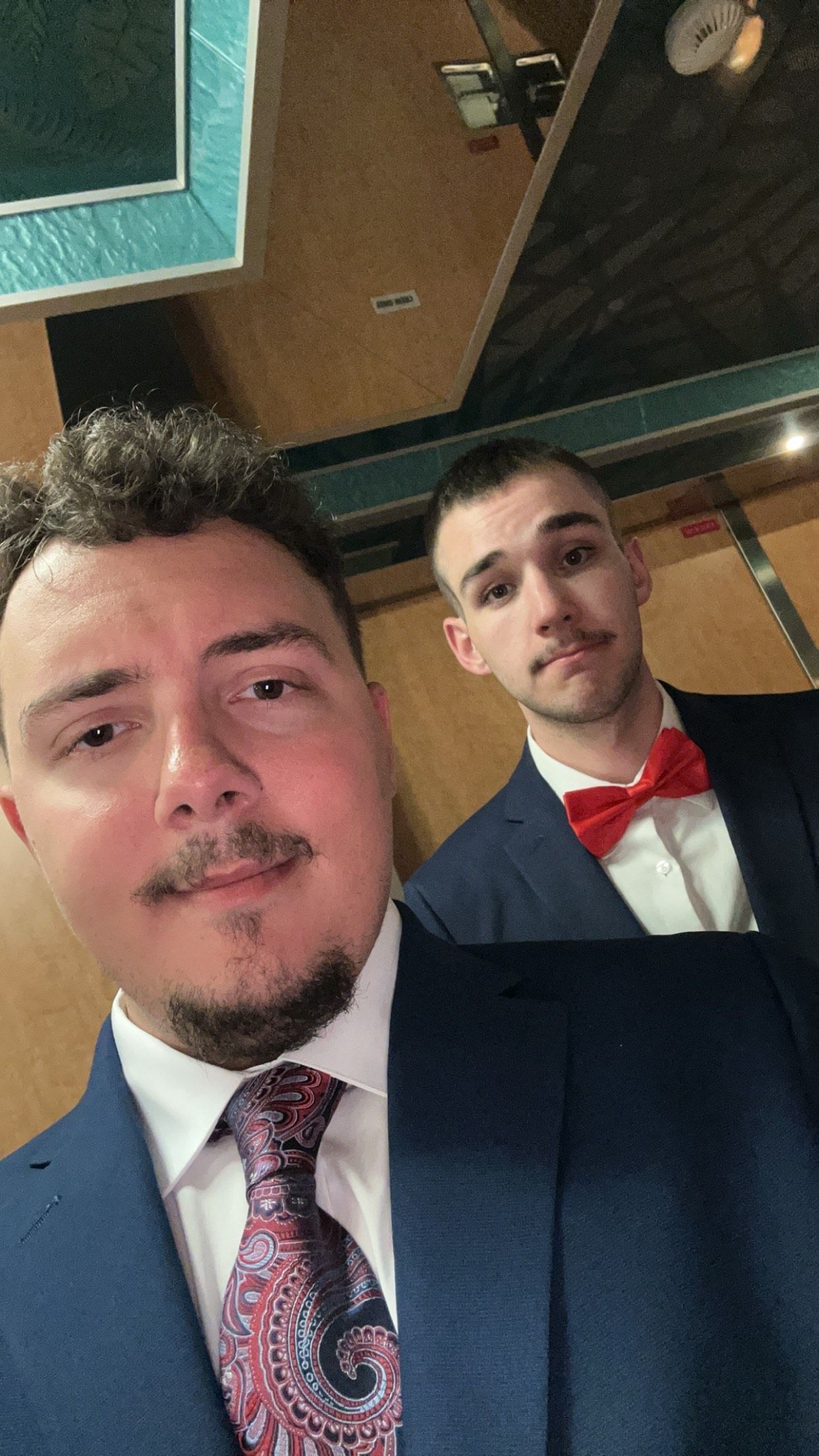 Two men in formal blue suits pose together, one wearing a red bow tie and one wearing a patterned necktie.