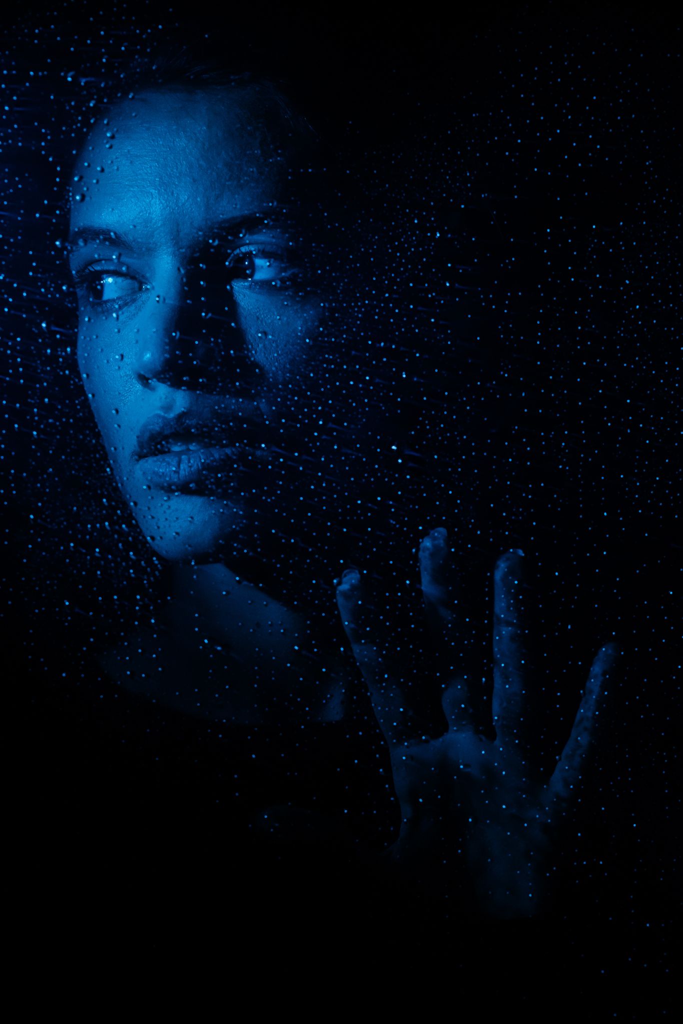 A dramatic portrait basked in deep blue light showing a silhouetted figure's face with a raised hand against a dark background.