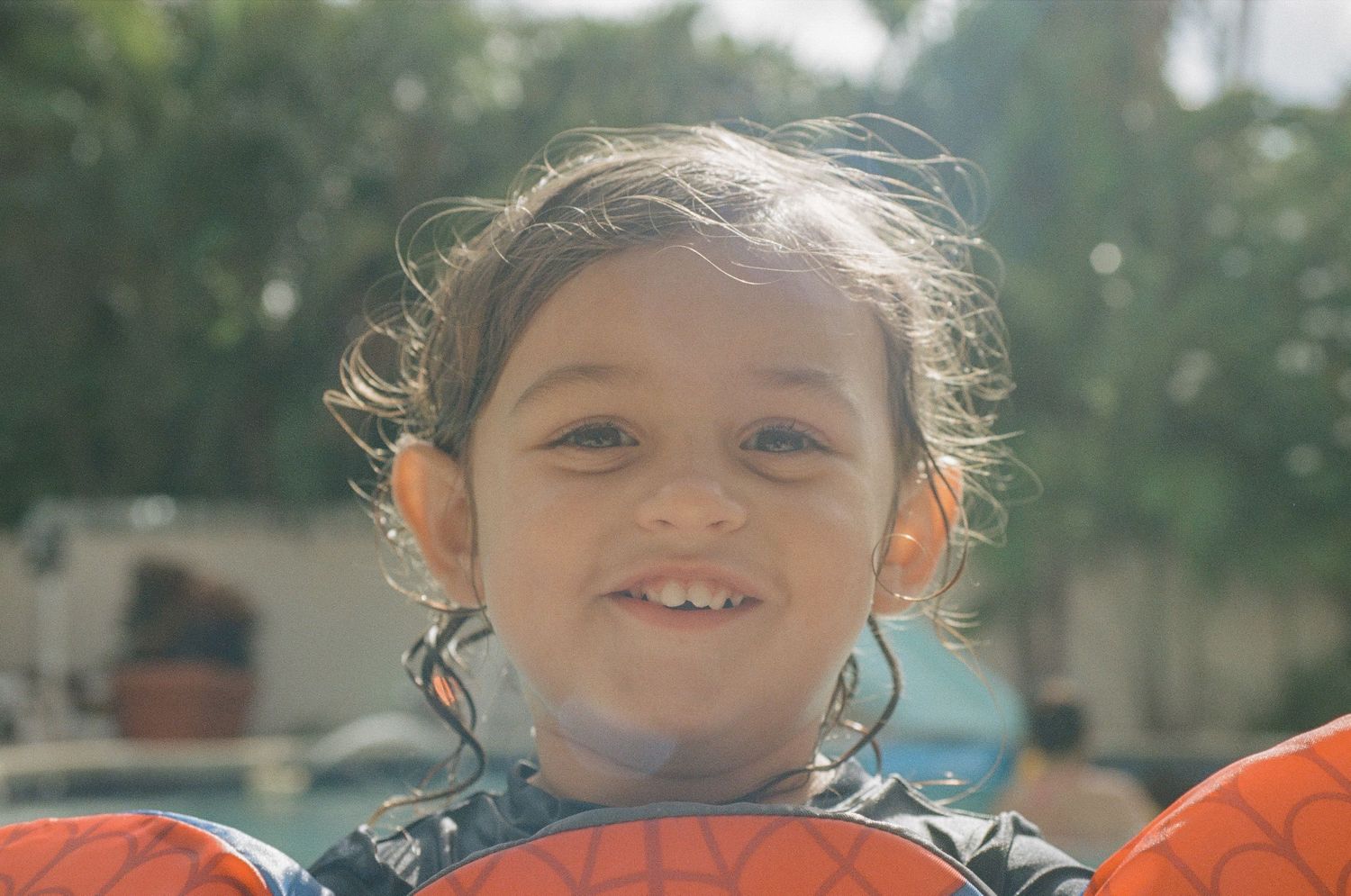 Closeup portrait of young child wearing orange life jacket with sunlight and blurred trees in background.